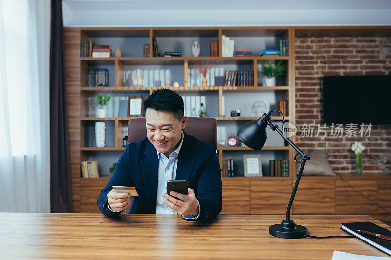 Young handsome asian man in suit in the office at work holding a credit card and shopping, ordering online from the phone, happy